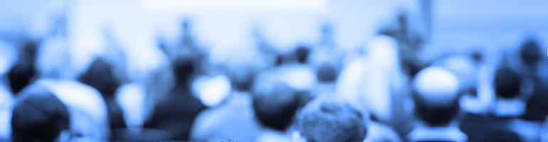 Back of a person's head in the foreground, sharply in focus, with an audience of blurred silhouettes seated in rows facing a large blank white screen or presentation board in a dimly lit conference or auditorium setting.