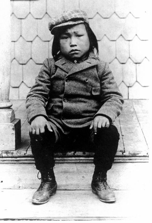 A black and white photo of a 7 year old Inuqhuaq named Mink from Greenland , sitting on a wooden porch in New York City. His hands are on his knees and he is dressed in American style clothes including a hat with flaps, a double-breasted overcoat and black pants. He was one of 6 Inuqhauq captured by the explorer Robert Peary and given to the American Museum of Natual History for anthropological and scientific study.