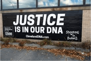 Photograph of a sign, affixed to the bottom wall of a building just beneath several reflecting windows. The sign is on a black background with white text that states "Justice In Our DNA." The sign is from an exhibit titled In "Our DNA: Arts, Activism and Black Cleveland " that was sponsored by the Department of Art and History at Case Western Reserve University in Cleveland, Ohio.